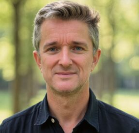 A Middle-Aged Man With Short, Graying Hair And Stubble Smiles Softly, Standing Outdoors In A Sunlit, Green, Blurred Park. He Wears A Black Collared Shirt. A Middle-Aged Man With Short, Graying Hair And Stubble Smiles Softly, Standing Outdoors In A Sunlit, Green, Blurred Park. He Wears A Black Collared Shirt.