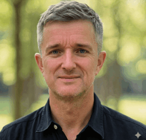 A Middle-Aged Man With Short Gray Hair And A Slight Smile Is Standing Outdoors, Wearing A Black Collared Shirt. The Background Is Blurred Greenery With Sunlight Filtering Through Trees.